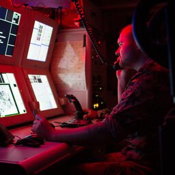 Ein Soldat sitzt in einem abgedunkelten, mit Rotlicht gefüllten, Raum und schaut auf Monitore