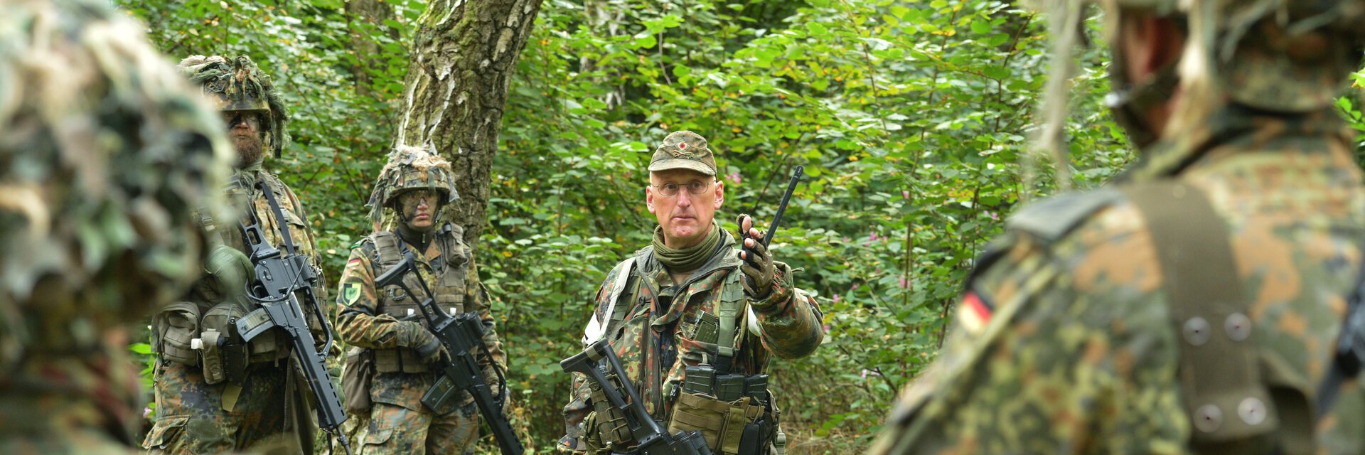 Soldaten in Flecktarnuniform mit Helm und Gewehr stehen in einem Wald und besprechen eine Lage