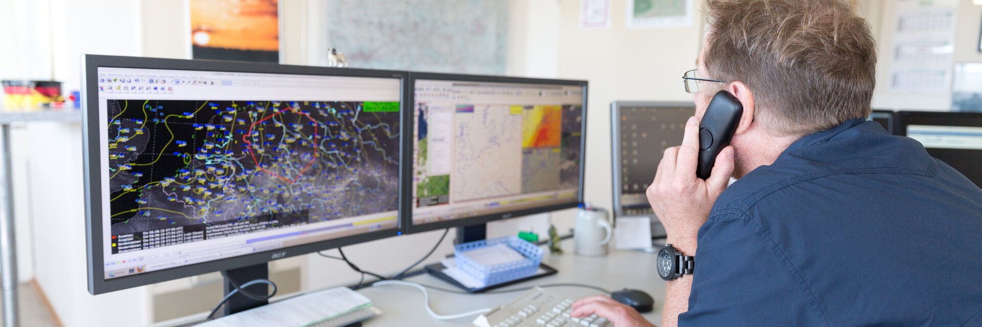 Ein Mitrbeiter des Wetterdienstes telefoniert und schaut dabei auf seine zwei Monitore mit Wetterkarten