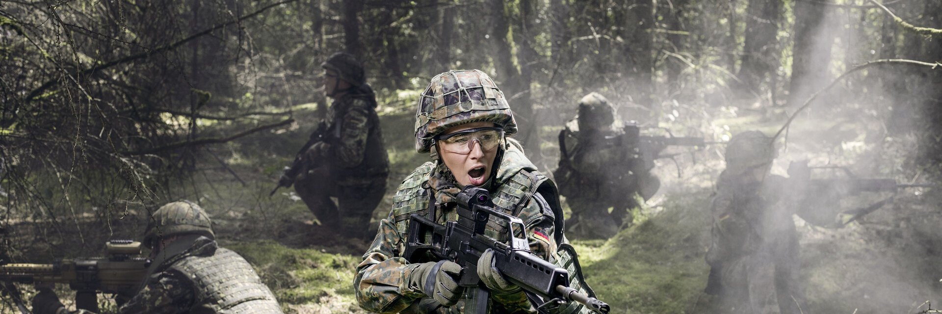 Soldatinnen und Soldaten liegen bei einer Ãœbung auf dem Waldboden und sichern mit Gewehren die Umgebung.
