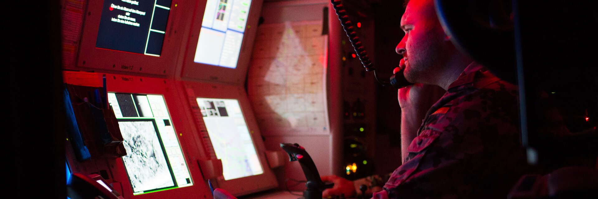 Ein Soldat sitzt in einem abgedunkelten, mit Rotlicht gefÃ¼llten, Raum und schaut auf Monitore