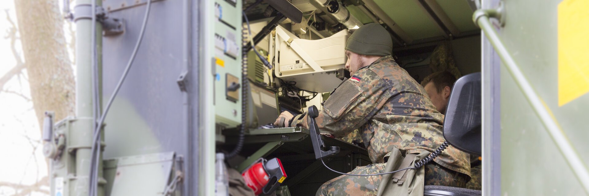 Ein Soldat in Uniform sitzt in einem Metallcontainer, der elektronisches Equipment enthÃ¤lt und schaut auf einen Monitor