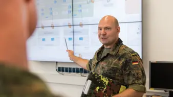 Ein Offizier in Uniform steht vor einer Tafel und unterrichtet andere Soldatinnen und Soldaten