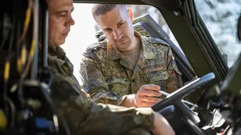 Ein Fahrlehrer in Uniform erklärt einem Fahrschüler, der in einem Bundeswehrfahrzeug auf dem Fahrersitz sitzt, die Anzeigen auf dem Armaturenbrett.