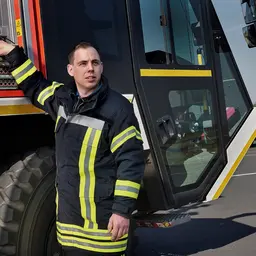 Ein Feuerwehrmann in Arbeitskleidung steh vor einem Löschfahrzeug der Flughafenfeuerwehr