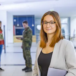 Eine Aufnahme in einer Universität. Im Vordergrund steht eine Frau und im Hintergrund unterhält sich ein Soldat mit einer zivilen Dame.