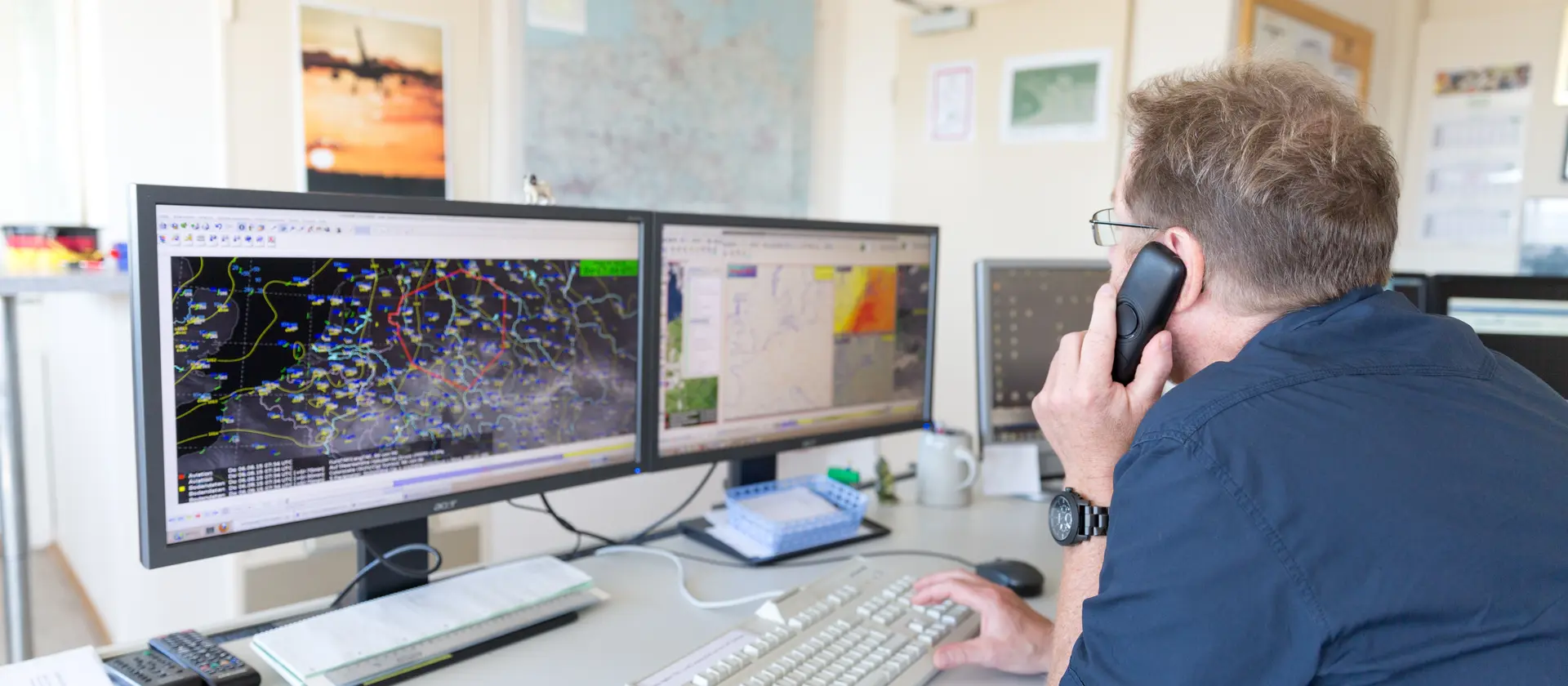 Ein Mann telefoniert und schaut dabei auf seine zwei Monitore mit Wetterkarten