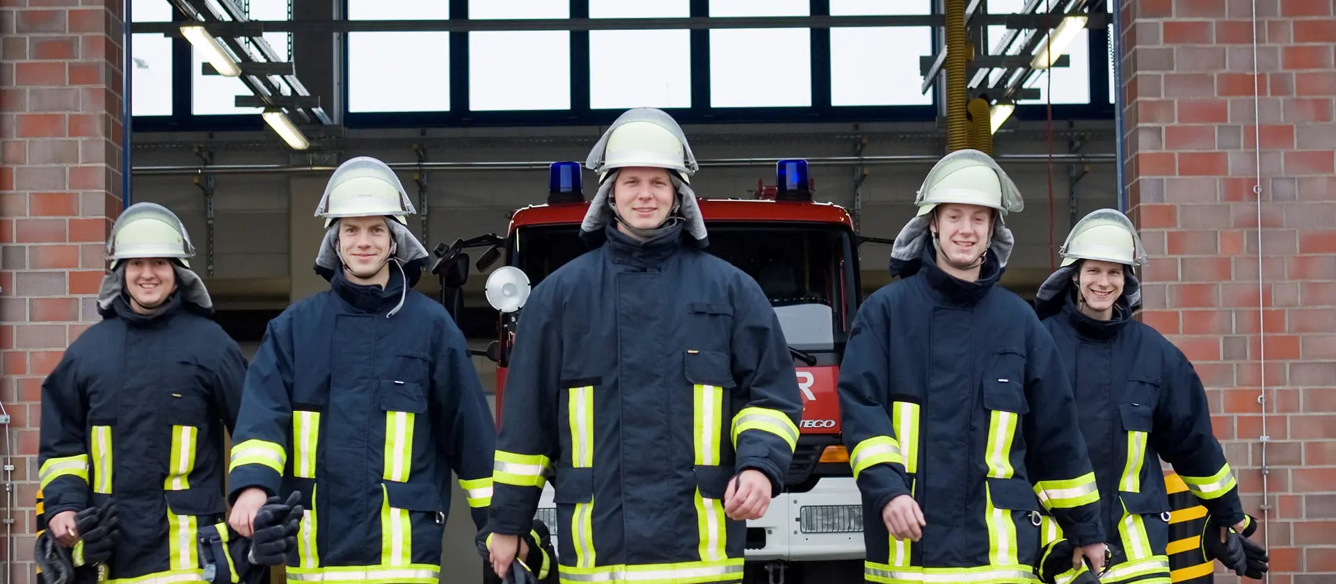 Eine Gruppe von fünf Feuerwehrleuten kommt nebeneinander aus einer Fahrzeughalle