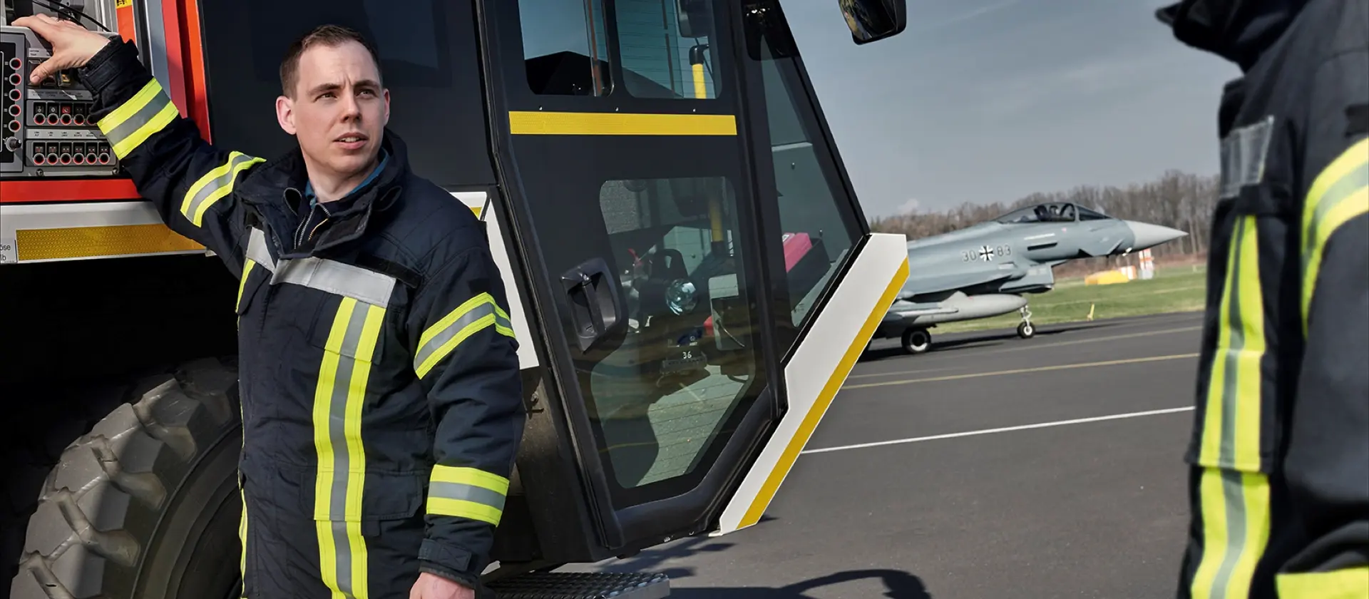 Ein Feuerwehrmann in Arbeitskleidung steh vor einem Löschfahrzeug der Flughafenfeuerwehr
