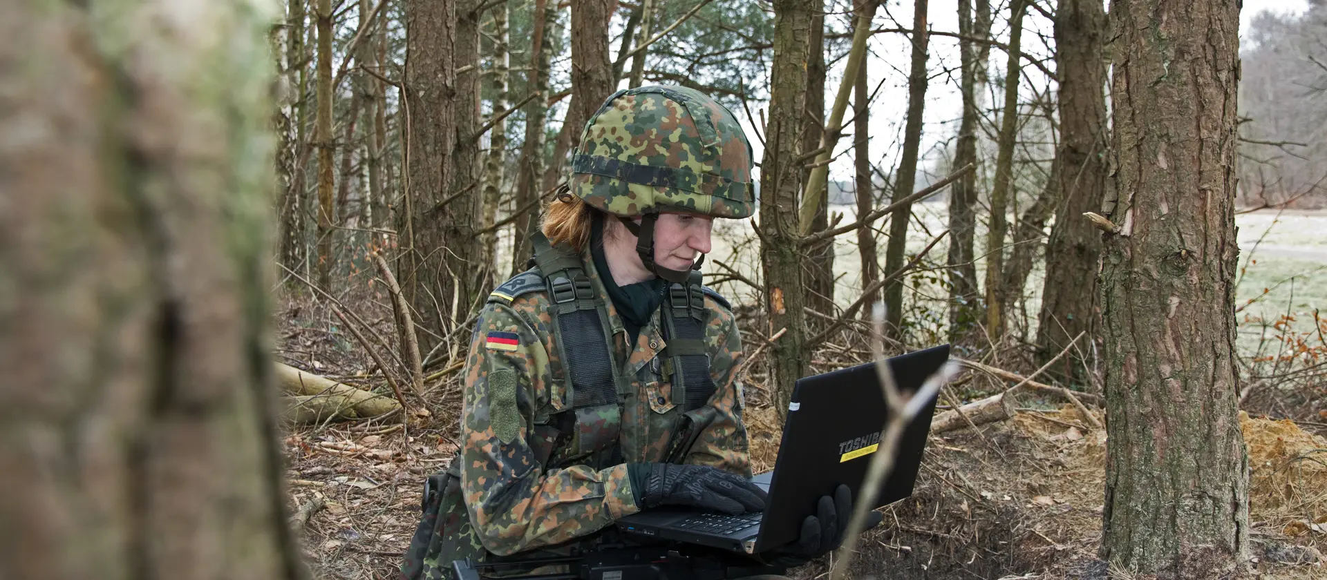 Eine Soldatin sitzt mit einem Laptop in einem Waldgebiet