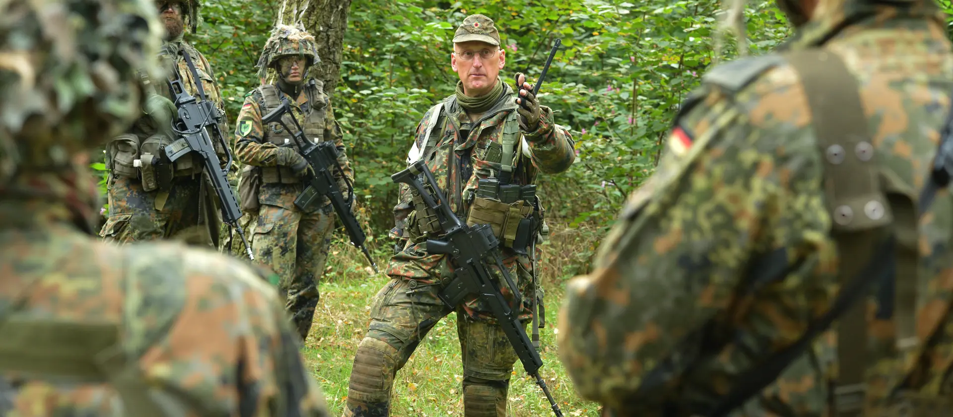 Soldaten in Flecktarnuniform mit Helm und Gewehr stehen in einem Wald und besprechen die Lage
