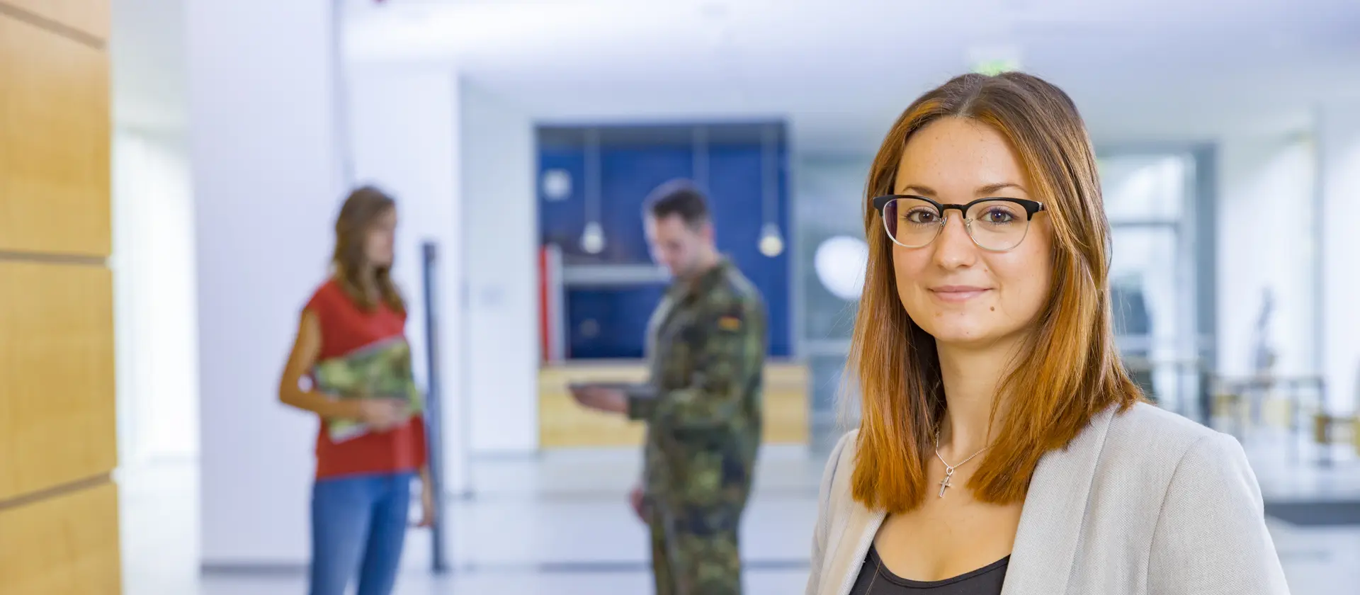 Eine Aufnahme in einer Universität. Im Vordergrund steht eine Frau und im Hintergrund unterhält sich ein Soldat mit einer zivilen Dame.