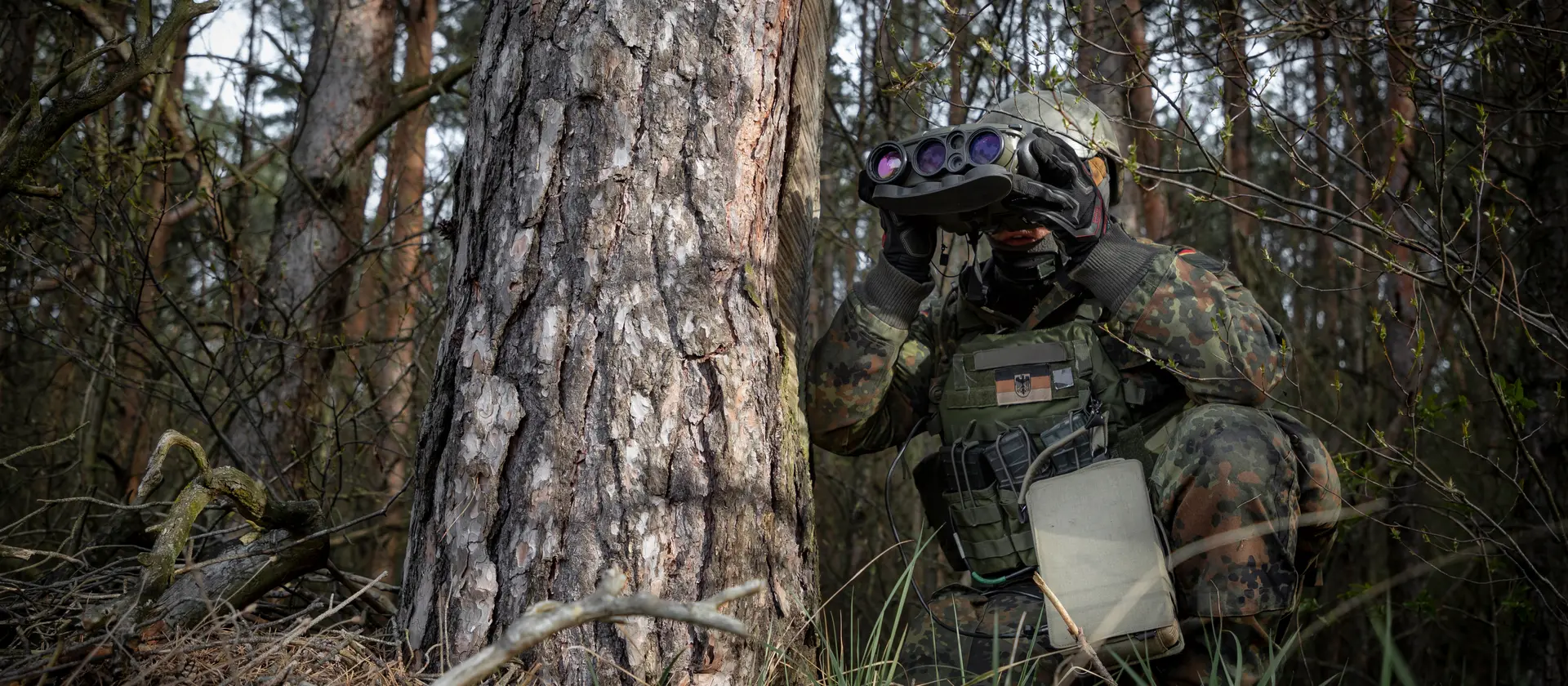 Ein Soldat in Flecktarnuniform sitzt neben einem Baum udn beobachtet den Luftverkehr als Koordinator für Luftnahunterstützung