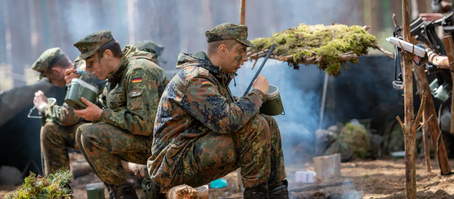Drei Soldaten in Uniform sitzen draußen im Gelände und essen etwas
