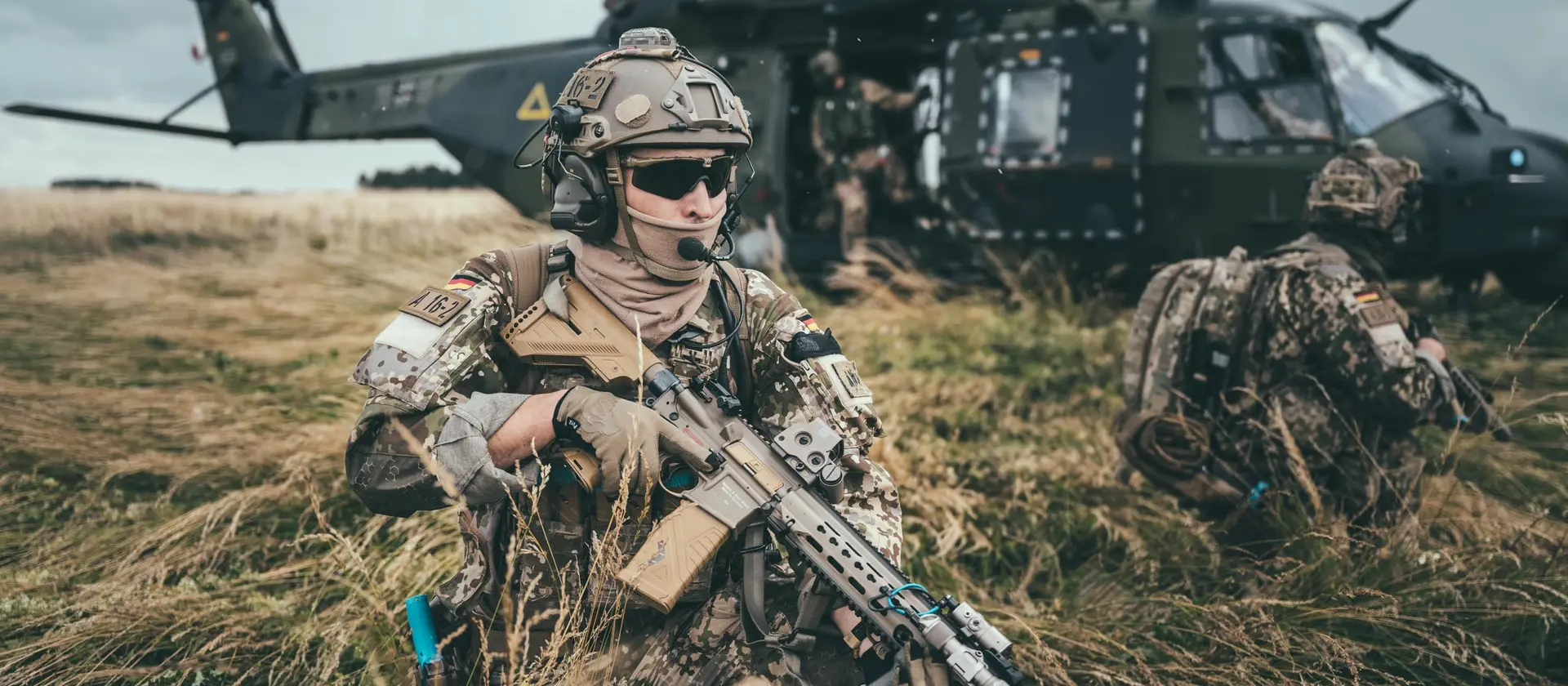 Soldaten abgehockt auf einem Feld vor einem Hubschrauber, sie tragen Gewehre und sichern die Umgebung