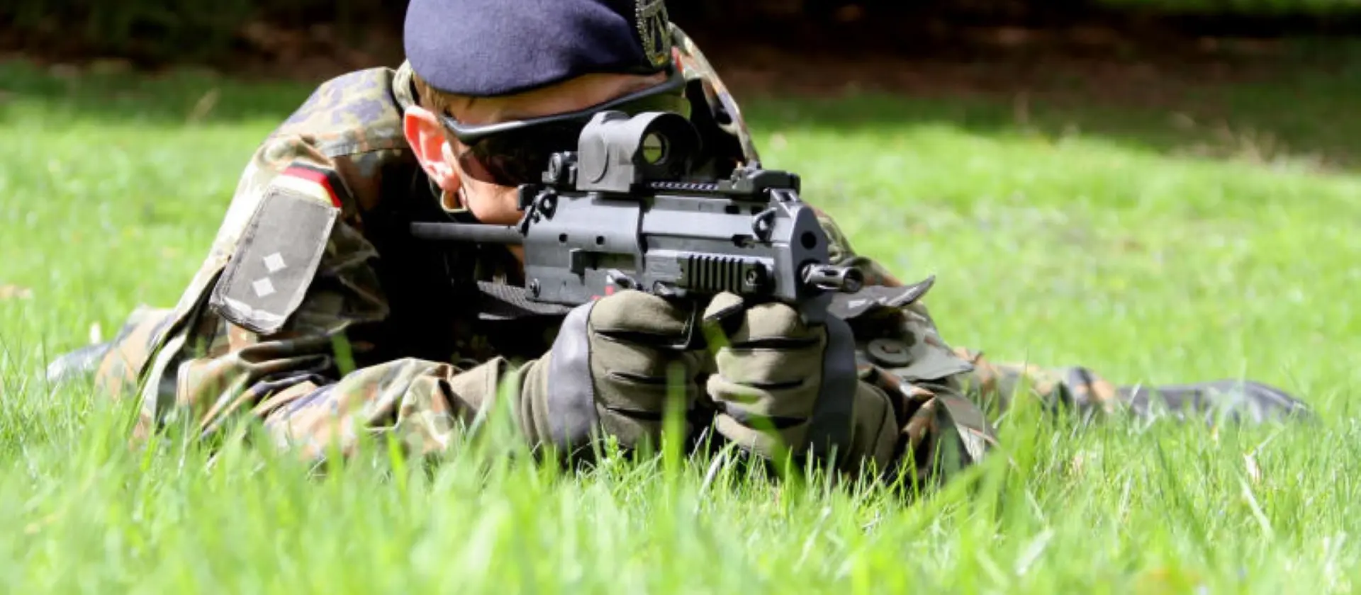 Sicherungssoldat mit Waffe liegt auf einer Wiese und beobachtet.