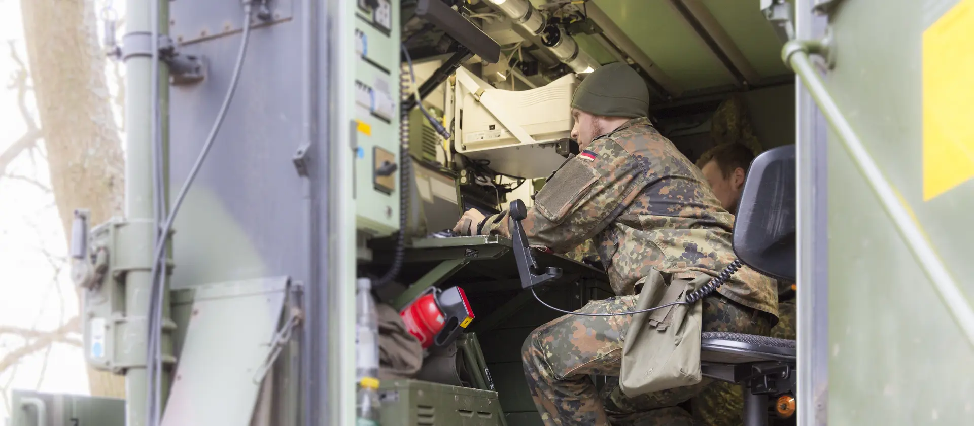 Ein Soldat in Uniform sitzt in einem Metallcontainer, der elektronisches Equipment enthält und schaut auf einen Monitor