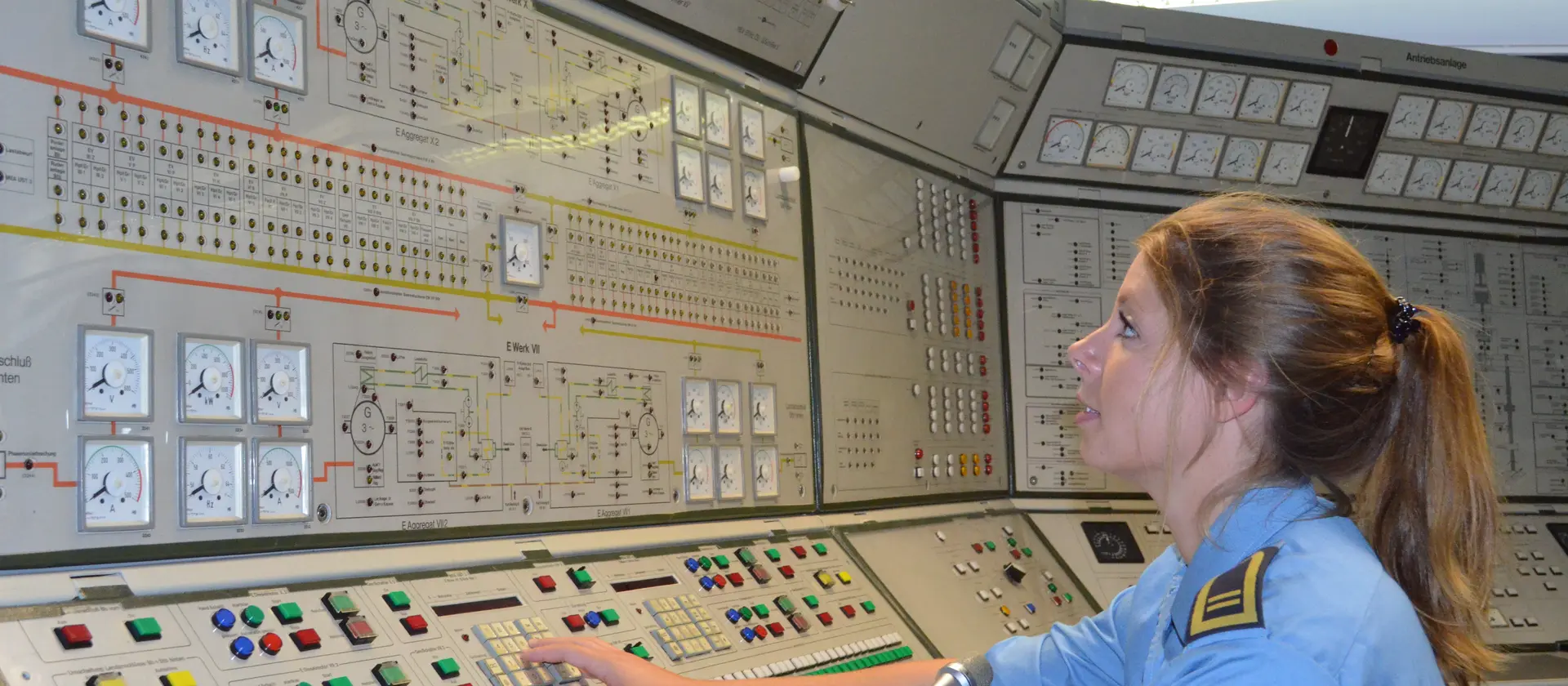 Eine Soldatin der Elektrotechnik der Marine sitzt vor einem großen Pult mit vielen Knöpfen und prüft technische Verbindungen.