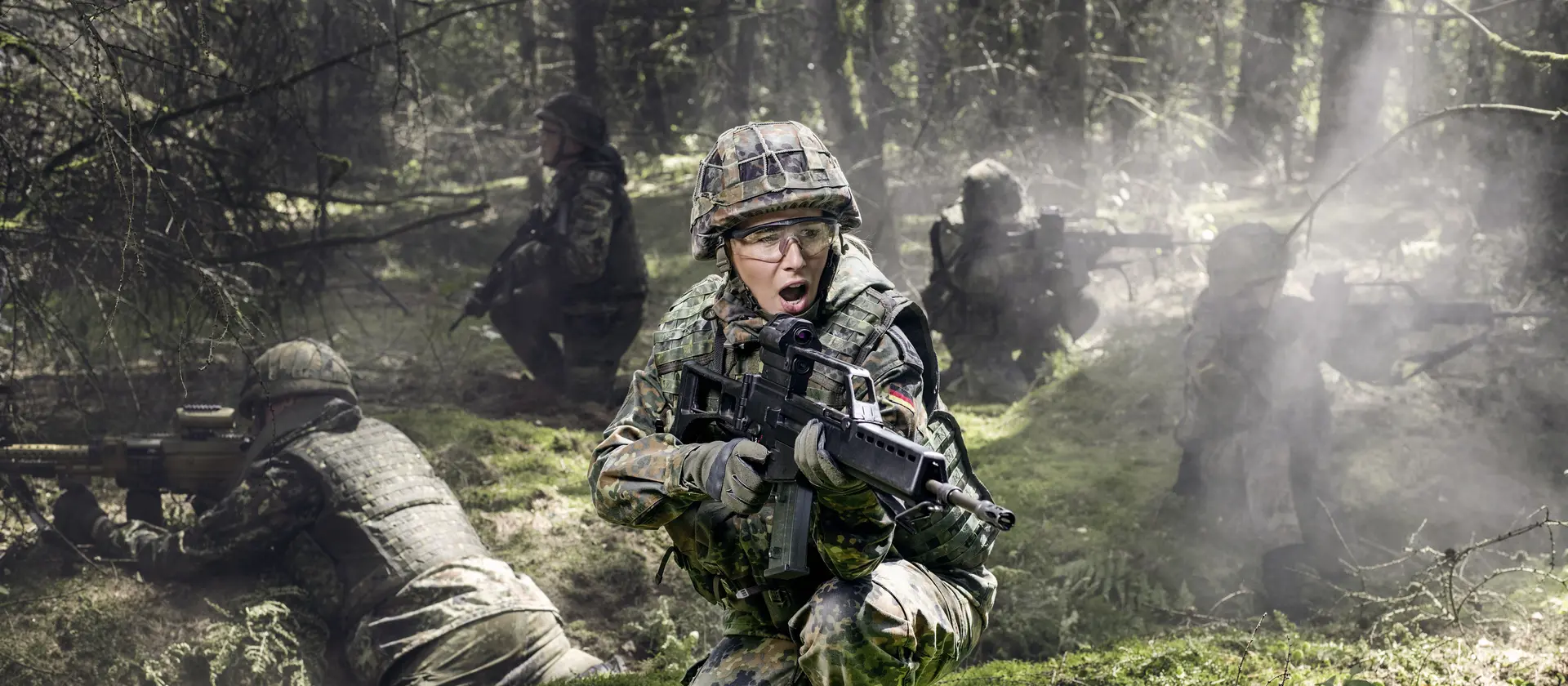 Soldatinnen und Soldaten liegen bei einer Übung auf dem Waldboden und sichern mit Gewehren die Umgebung.