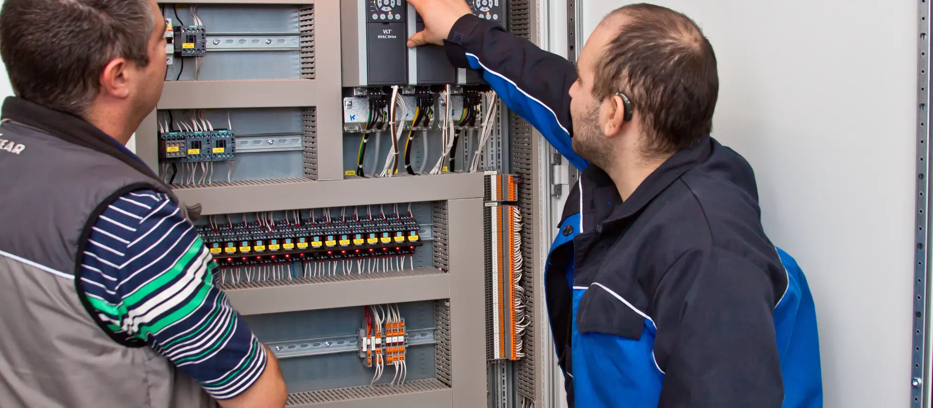 Zwei Herren stehen nebeneinander und schauen auf einen elektronischen Schaltkasten. Hierbei wird gerade ein Wert auf einem Display abgelesen.