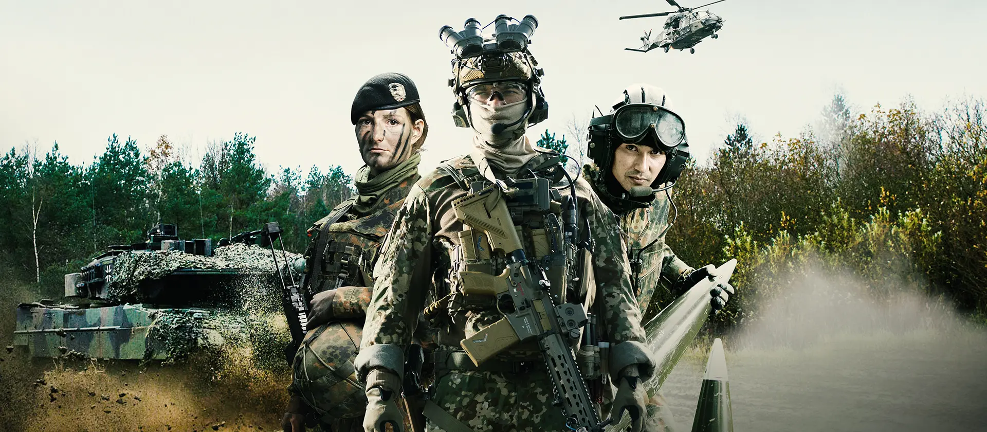 Eine Soldatin und zwei Soldaten in Kampfmontur stehen bildmittig mit Blick in die Kamera auf einem Feld. Im Hintergrund ist ein Wäldchen zu sehen. Links der Gruppe von Soldat/innen ist ein Schützenpanzer, über der Gruppe ein Bundeswehrhubschrauber zu sehen.