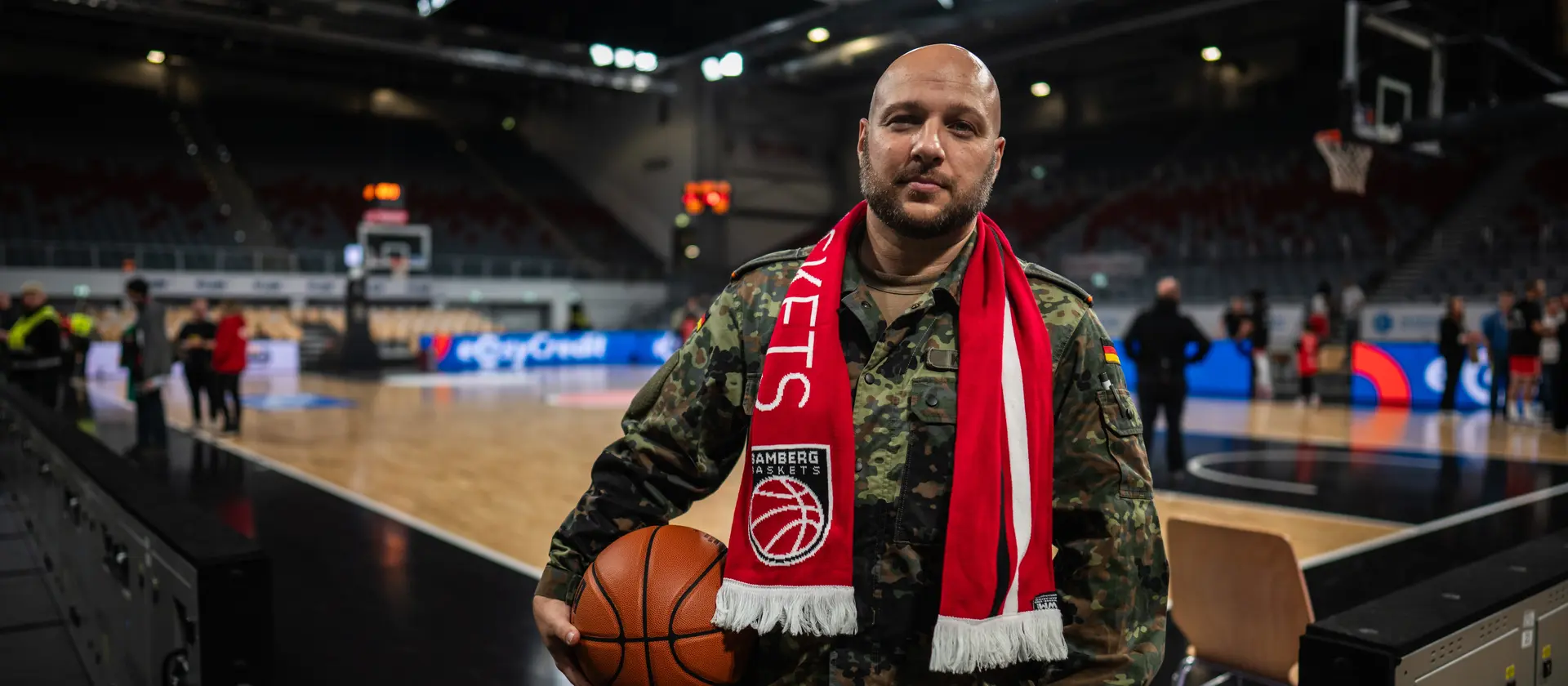 Ein Soldat der Bundeswehr steht mit einem Basketball und einem Schaal vor einem Basketballfeld