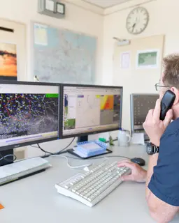 Ein Mann telefoniert und schaut dabei auf seine zwei Monitore mit Wetterkarten